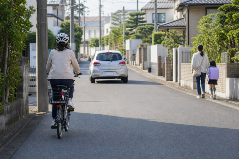 中央線のない日本の生活道路を左側通行するヘルメット着用の自転車と、間隔を空けて走る自動車、道路脇を歩く親子