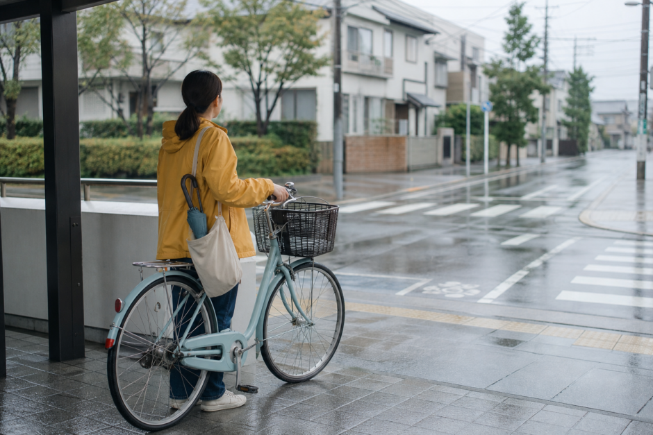 雨の日に傘を差さずレインウェアで自転車のそばに立ち周囲を確認する利用者