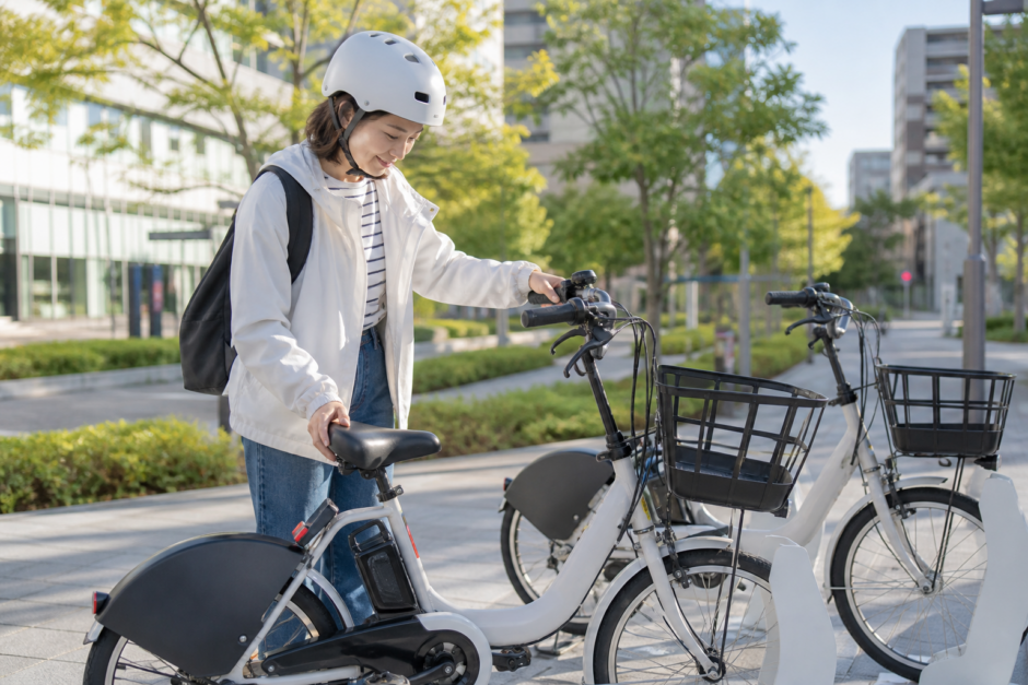 街角のシェアサイクルポートでヘルメットを着用した利用者が電動アシスト自転車のサドルを確認しているイメージ