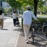夏の明るい街路でヘルメットを着用した自転車利用者が歩行者に配慮しながら駐輪場へ向かう安全啓発イメージ
