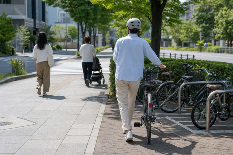 夏の明るい街路でヘルメットを着用した自転車利用者が歩行者に配慮しながら駐輪場へ向かう安全啓発イメージ