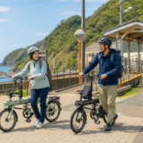 海辺の小さな駅前でヘルメットを着用した大人が折りたたみ自転車を押している明るい旅行風景