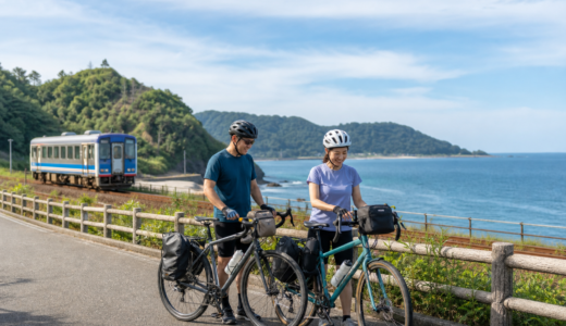 日本海沿いの道でヘルメットを着用したサイクリストが自転車を押し、列車と海を背景に旅の準備をしているイメージ