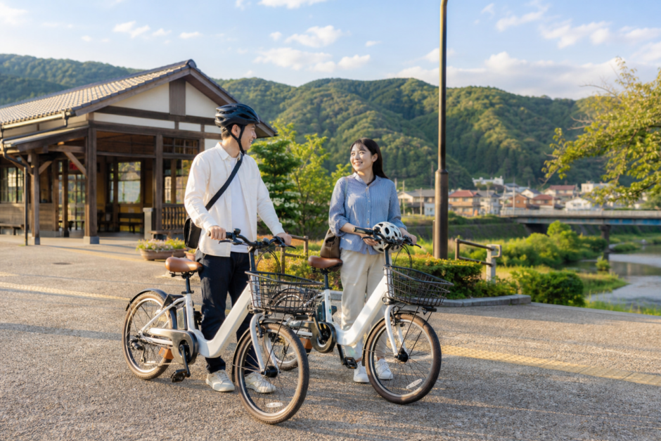 雲南市の里山に近い駅前でヘルメットを持った大人がシェアサイクルの電動アシスト自転車を準備しているイメージ