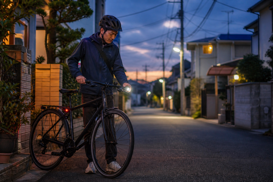 夕暮れの住宅街でヘルメットを着用した自転車利用者が前照灯の点灯を確認している様子