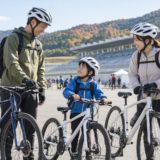 鈴鹿サーキットのような広い会場でヘルメットを着用した親子とサイクリストが自転車イベント前に安全確認をしている様子